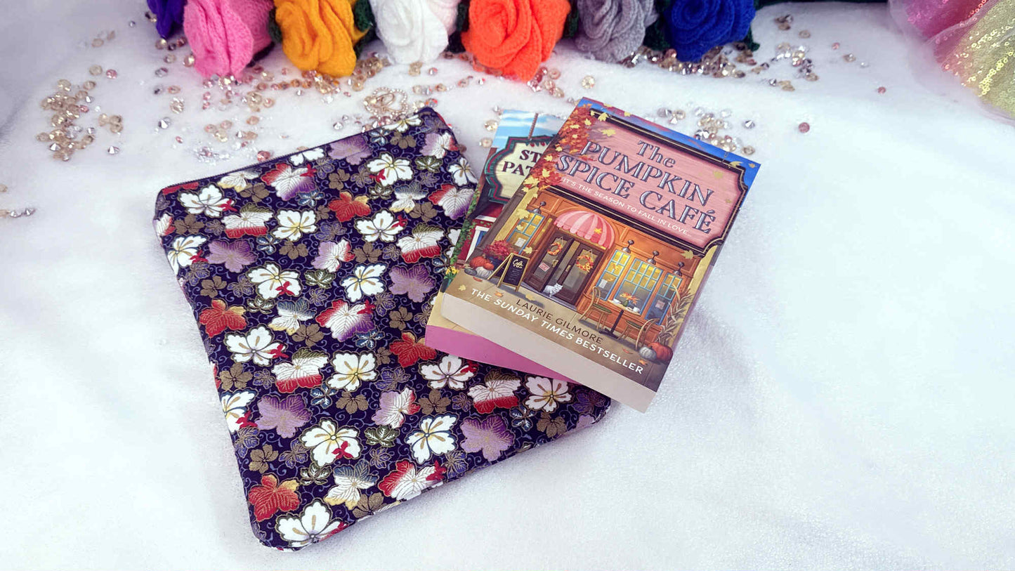 Floral-patterned pouch and book titled 'The Pumpkin Spice Cafe' on a white surface with colorful yarn balls in the background. Handmade book sleeve by Roxana Roveno